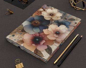 May include: A hardcover journal featuring a floral design with watercolor-style flowers in shades of blue, peach, and cream. The journal is rectangular and lies on a dark gray surface, with gold and black pencils and gold binder clips nearby.
