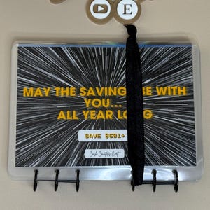 Puede incluir: Un archivador de presupuesto de plástico transparente con un diseño gráfico en blanco y negro y el texto "MAY THE SAVING BE WITH YOU... ALL YEAR LONG." El archivador tiene un cierre elástico negro y una columna vertebral negra con ganchos.
