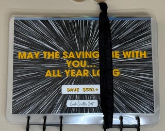 Pode incluir: Um fichário de orçamento de plástico transparente com um design gráfico em preto e branco e o texto "MAY THE SAVING BE WITH YOU... ALL YEAR LONG." O fichário tem um fecho elástico preto e uma lombada preta com ganchos.
