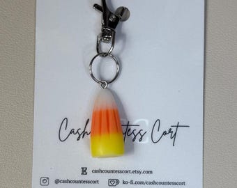 Pode incluir: Um chaveiro em forma de doce de milho com camadas brancas, laranja e amarelas. O pingente é preso a um mosquetão e um chaveiro de metal prateado. O fundo é um cartão branco com o texto "Cash Countess Cort".