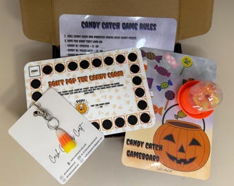 Pode incluir: Um conjunto de jogos com tema de Halloween com um chaveiro de milho doce, regras do jogo e tabuleiros. Os tabuleiros apresentam uma abóbora e o texto "Candy Catch Gameboard" e "Don't Pop the Candy Corn!"