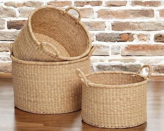 May include: Three round woven storage baskets in different sizes. The baskets have handles and a natural tan hue. The largest basket is at the back, with two smaller ones in front. They are on a wooden surface.