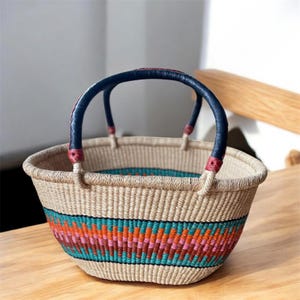 Puede incluir: Cesta tejida con un colorido patrón geométrico en turquesa, naranja y rosa. La cesta tiene un asa resistente con un agarre azul oscuro y detalles en rojo. La cesta está hecha de fibras naturales.