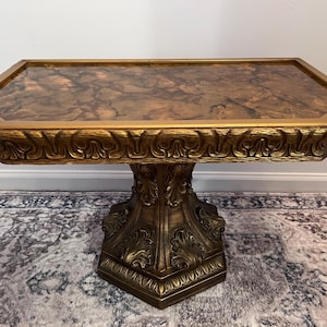 May include: Ornate gold-colored accent table with a hexagonal top and a detailed, carved base. The tabletop features a brown and gold marbled pattern. The table is approximately 30 inches long and 18 inches wide.