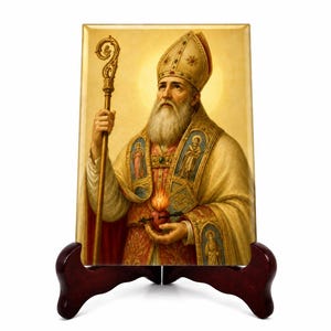 May include: A rectangular plaque featuring a religious painting of a bearded bishop in ornate gold and red vestments, wearing a mitre, holding a golden crozier, and cradling a flaming heart pierced by an arrow, displayed on a dark wooden stand.
