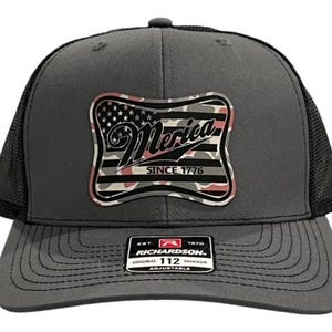 Może przedstawiać: Szara i czarna czapka trucker z naszywką w kamuflażu z amerykańską flagą, na której widnieje napis "Merica Since 1776". Czapka jest regulowana i ma siateczkowe tylne panele.