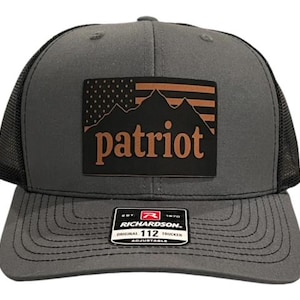 Può includere: Un cappello da camionista grigio e nero con una toppa marrone che dice "patriot" e presenta una catena montuosa e una bandiera americana.