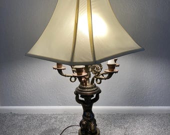 May include: A gold-toned table lamp with a white lampshade. The lamp base features a sculpted figure holding up a candelabra with five candle holders.