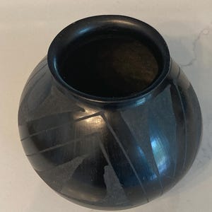 May include: A black ceramic pot with a rounded body and a wide opening. The pot features a matte finish and is decorated with etched geometric patterns. The design includes lines and triangular shapes. The pot is likely handmade.