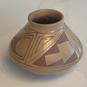 May include: A tan-colored ceramic pot with a rounded body and a narrow opening. The pot is decorated with geometric designs in dark brown and reddish-brown. The design includes triangles and angular shapes. The pot has a smooth, matte finish.