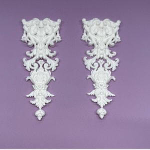 May include: Two white, ornate decorative appliques with a symmetrical design. Each applique features a detailed floral and scrollwork pattern, with a central floral element and a teardrop-shaped base. The appliques are set against a lavender background.
