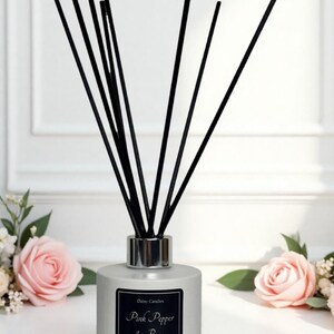 May include: A grey diffuser with black reeds and a silver collar. The black label reads "Pink Pepper & Rose". The diffuser is surrounded by pink roses and greenery, creating a floral and elegant aesthetic.