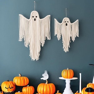 Könnte beinhalten: Eine Halloween-Deko mit zwei weißen Makramee-Geister-Wandbehängen, einem weißen Keramik-Geist mit einem schwarzen Hexenhut und einer Auswahl an orangefarbenen Kürbissen.