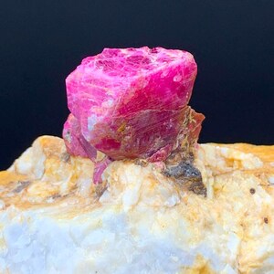 Puede incluir: Un espécimen de cristal de rubí rosa intenso incrustado en una matriz rugosa y texturizada de cuarzo blanco y naranja pálido. La piedra preciosa presenta una estructura bruta con facetas e inclusiones naturales visibles en su superficie.