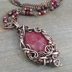 May include: A handcrafted copper pendant necklace with a faceted, deep red gemstone. The gemstone is encased in intricate wirework with small, decorative beads. The necklace has a copper chain with additional red beads.