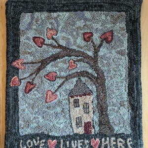 May include: A hand-stitched rug with a whimsical design of a house with a tree with hearts for leaves. The text "Love Lives Here" is stitched at the bottom of the rug.