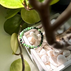 May include: A bracelet made of green and black beads, with small red and pink accents. The bracelet is presented on white pebbles within a white wooden tray, with green leaves in the background.
