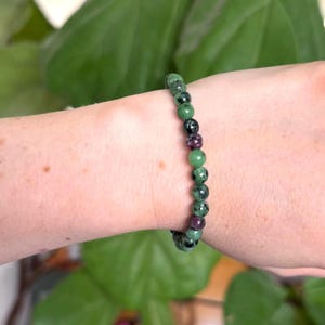 May include: A bracelet made of green, black, and maroon gemstone beads. The beads are small and round, with a speckled pattern. The bracelet is worn on a wrist. The background features green foliage. The bracelet is a piece of jewellery.