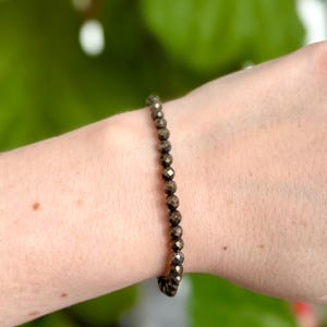 Peut inclure: Un bracelet en perles dorées composé de petites perles de pyrite facettées. Le bracelet est porté au poignet.