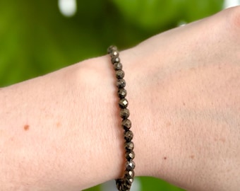 May include: A gold-toned beaded bracelet made of small, faceted pyrite beads. The bracelet is worn on a person's wrist.