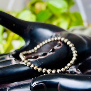 Puede incluir: Un brazalete de cuentas de pirita de color dorado con acabado facetado. El brazalete está apoyado en una mano negra.