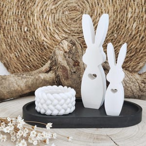 Könnte beinhalten: Weiße Osterhasen-Figuren mit Herz-Ausschnitten, ein weißer geflochtener Kerzenhalter und ein schwarzes Tablett. Die Anordnung befindet sich auf einer Holzoberfläche, mit einem natürlichen Webhintergrund.