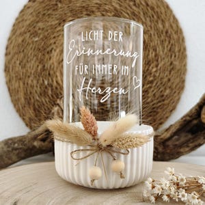 Könnte beinhalten: Ein klarer Glaszylinder mit dem weißen Schriftzug "LICHT DER ERINNERUNG FÜR IMMER IM HERZEN" und einem kleinen Herzsymbol. Das Glas steht in einer weißen, gerippten Keramikschale, die mit getrockneten Gräsern, Kordel und zwei Holzperlen verziert ist.