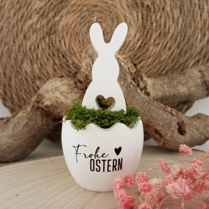 Könnte beinhalten: Weißer Keramik-Oster-Eiertopf mit einer Hasensilhouette und dem Text "Frohe Ostern" in schwarzer Schrift. Der Topf ist mit grünem Moos gefüllt.