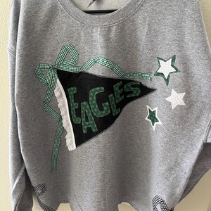 May include: Gray sweatshirt with a black and green pennant that says "EAGLES" in white letters. The pennant is decorated with green and white gingham ribbon and white stars.