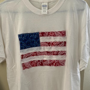 May include: White t-shirt with a red, white, and blue American flag design made from fabric scraps and lace. The flag is centered on the shirt.