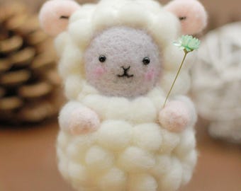 Pode incluir: Um pequeno brinquedo de ovelha de feltro branco com um rosto roxo claro e orelhas cor-de-rosa. A ovelha está segurando uma pequena flor verde.