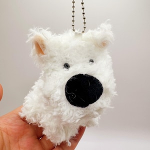 Op de afbeelding: Witte pluche honden sleutelhanger met een zwarte neus. De sleutelhanger is bevestigd aan een zilveren ketting.