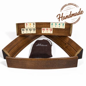 May include: A set of four curved wooden racks for a tile game, featuring dark brown wood grain and brass-colored metal accents labeled HELENA. A brown drawstring pouch with the text Helena Woodart is centered among the racks.