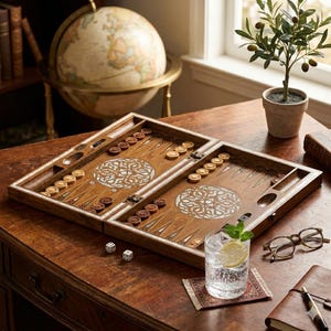 May include: A wooden backgammon set with light and dark brown checkers and dice sits on a wooden table. A globe, a small potted olive tree, and a glass of water with a lemon slice are also on the table.