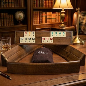May include: A wooden Rummikub game set with tiles, racks, and a carrying bag. The tiles are white with black and red numbers. The set is on a brown leather surface, with a pen and glasses visible.