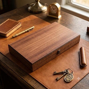 May include: A rectangular wooden storage box with a visible grain pattern and a small metal latch closure. The box is positioned on a brown leather desk mat alongside a gold-toned pen, a small leather-bound notebook, a clock, and a metal keychain.