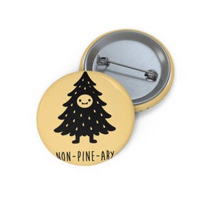 May include: Two round, yellow pinback buttons. One button features a black cartoon Christmas tree with a smiling face and the text "NON-PINE-ARY". The other button is a silver metal backing.