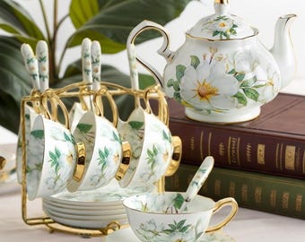 May include: A white porcelain tea set with a floral design. The set includes a teapot, six cups, six saucers, and a stand. The teapot has a gold trim and a floral design with white flowers and green leaves. The cups and saucers also have a gold trim and a floral design.