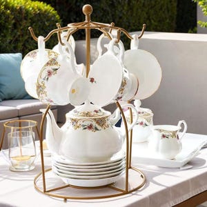 Puede incluir: Un soporte de metal dorado para tazas y platillos de té con una tetera, una jarra de crema, un azucarero y seis tazas y platillos de té. Las tazas y platillos tienen un patrón floral con acentos dorados.