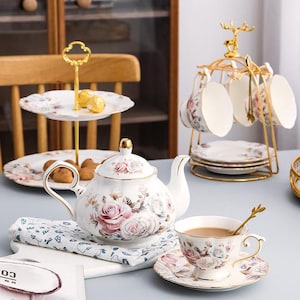 May include: A white porcelain teapot with a floral design and a gold handle. The teapot is sitting on a white table with a white teacup and saucer, a two-tiered serving stand, and a gold metal teacup holder.