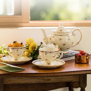 Könnte beinhalten: Ein Teeservice im Vintage-Stil mit einer weißen Teekanne und zwei Teetassen mit Blumenmustern. Die Teekanne hat einen Deckel und einen Henkel, die Teetassen haben Untertassen. Das Teeservice steht auf einem Holztisch mit einem grünen Umschlag und einem kleinen Kupfertopf mit roten Tomaten.