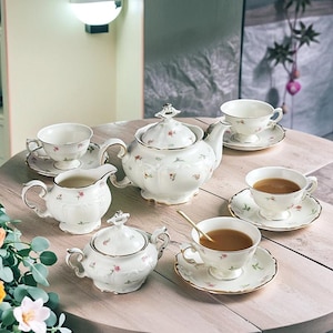 Puede incluir: Un juego de té de porcelana blanca con diseños florales. El juego incluye una tetera, azucarero, lechera y varias tazas de té con platillos. Las tazas de té están llenas de té.
