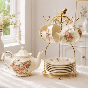 Puede incluir: Un juego de té de color crema que incluye una tetera y varias tazas con motivos de rosas rosadas y bordes dorados. Las tazas y los platillos están organizados en un soporte de metal dorado rematado con una pequeña figura de pájaro.