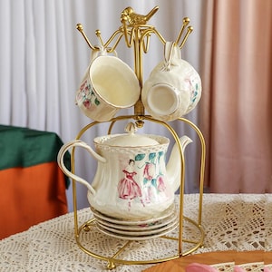 May include: A gold-colored metal tea set stand with a teapot, cups, and saucers. The teapot and cups are cream-colored with floral and ballerina designs. A small gold bird sits atop the stand. The set is arranged on a lace tablecloth.