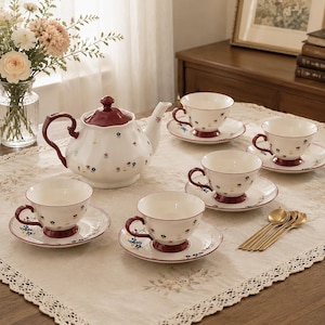Puede incluir: Un juego de té de cerámica blanca que incluye una tetera y cuatro tazas con sus platos a juego, decorados con pequeños motivos florales coloridos y detalles en rojo oscuro en las asas, tapas y bases, junto con cuatro cucharas de metal dorado.
