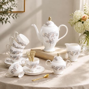 May include: A white porcelain tea set featuring a delicate grey hydrangea floral pattern and gold trim. The set includes a teapot, a lidded sugar bowl, a creamer, six teacups with matching saucers, and several ornate gold-toned metal spoons.