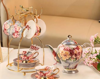 May include: A floral tea set with a teapot, cups, saucers, and a gold metal stand. The teapot and cups feature a floral design with roses and other flowers in shades of pink, yellow, and red. The stand has a bird design on top.