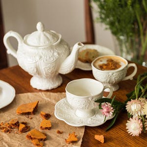 Op de afbeelding: Een wit porseleinen theeservies met reliëf bloemmotieven. De set bevat een theepot, kopjes, schotels en een kopje koffie. De scène speelt zich af op een houten tafel met een snack en bloemen.