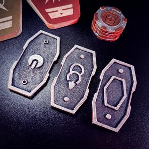 May include: Three silver metal game pieces with different symbols. The pieces are shaped like shields and have a textured surface.