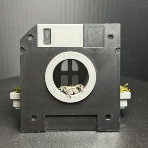 May include: A black 3D-printed floppy disk enclosure featuring a circular white window with a black arched frame. A small, fuzzy, light-colored jumping spider is positioned inside the circular opening, with green moss visible on the sides.